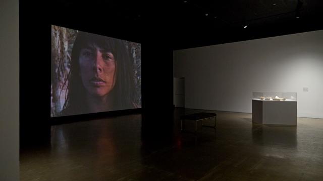 A dark gallery has a diffuse spotlight on an artistic stand, a screen in the background shows the face of an indigenous woman.