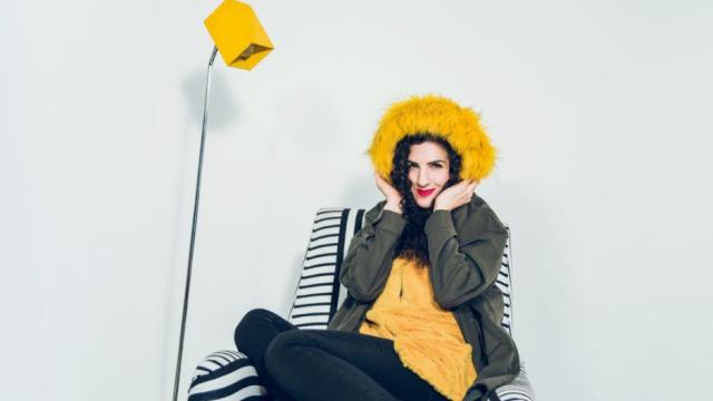 Laila Biali is wearing a bright yellow shirt and matching fur hat, while sitting on a black and white striped armchair, next to a lamp with a yellow lampshade.