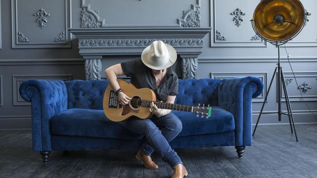 Irish sits on a blue felt couch wearing a broad rimmed hat playing her guitar.