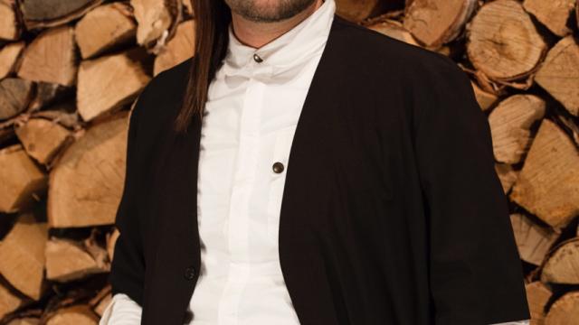 Visual Artist John G. Hampton stands in front of a wall of split logs.