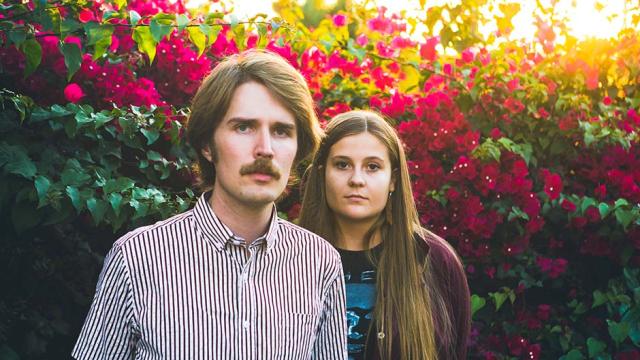 Kacy and Clayton are standing outdoors in front of a colorful hedge of green leaves and fuschia flowers. 