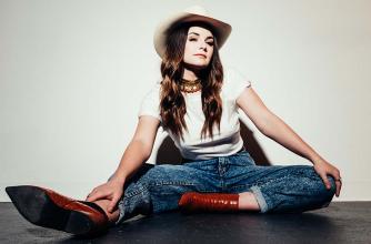 Mariya Stokes is wearing brown boots, blue jeans, white t-shirt, and a white cowboy hat. She is seated on the ground in front of a white wall. 