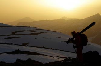 Silhouetted skier with skis resting on shoulder stands against a backdrop of snow-covered mountains as the setting sun casts a warm glow, creating a dramatic alpine scene