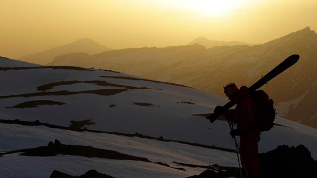 Silhouetted skier with skis resting on shoulder stands against a backdrop of snow-covered mountains as the setting sun casts a warm glow, creating a dramatic alpine scene