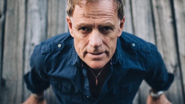 Martyn Joseph sits flat on a deck with his bands behind him propping himself up and looking at the camera.