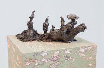 Cold-cast aluminum, resin, soil, rocks, 5 x 14 x 6 inches. The sculpture appears root-like with different protrusions, it is seated atop a faded green and silver burnished cube.