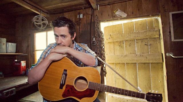 Danny Michel is seated with both arms resting on the guitar on his lap