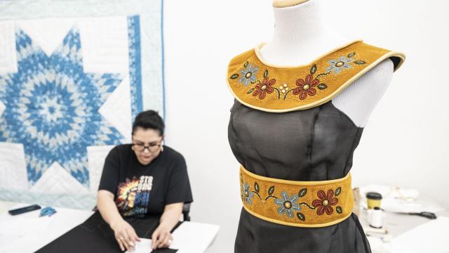 An artist is seated in a studio next to a black embellished dress on a dress form. 