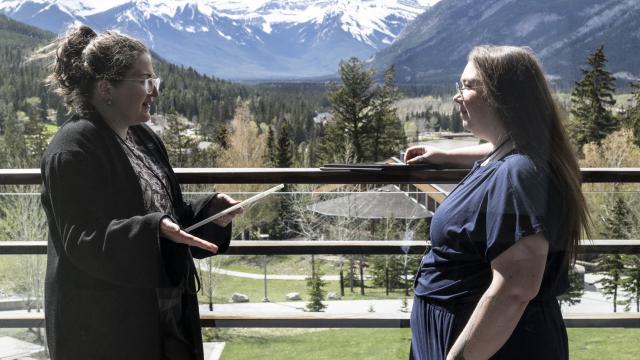 Two people are standing outdoors with mountains in the background. They are having a conversation. 