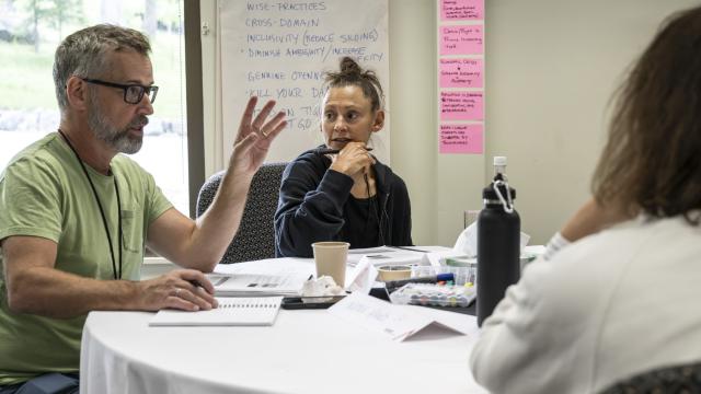Three people are seated at a conference table. One of them is speaking while the others listen.