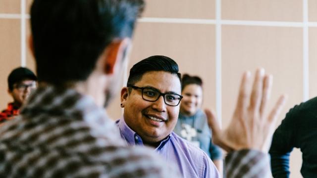 Photo shows Students in Introduction Negotiation Skills Training Course, one student stands to left with back to camera and right hand up and open as an another student looks forward to high five.