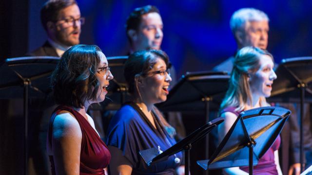 Six choral singers are standing in two rows of three and singing to an audience off to the right of the camera