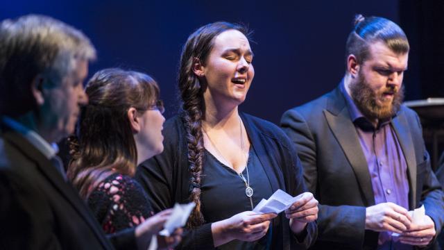 A group of four people stand on stage. A woman with a long braid, centered, sings wtih her eyes closed. A bearded man in a blazer stands to her right.