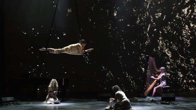 Aerial performer suspended on a trapeze bar, flying horizontally above a dimly lit stage with a backdrop of scattered golden light particles. Below, two performers kneel with metal bowls, and a harpist in white plays a concert harp at right; a piano sits in shadow at left.