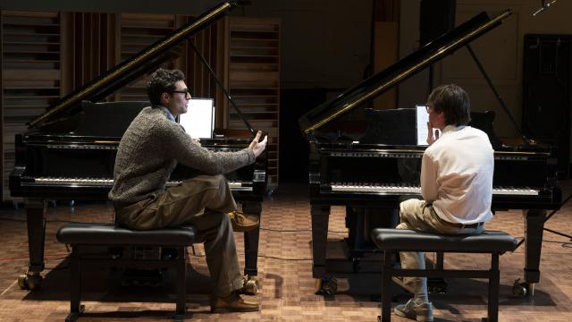 A teacher and a student are seated at side by side grand pianos, with the teacher instructing the student