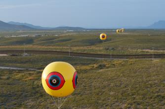 Yellow helium balloons with an eye symbol are spread out across an open landscape