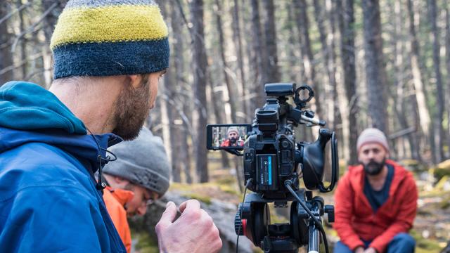 Person wearing a blue jacket and striped hat points video camera at man in red jacket, seated