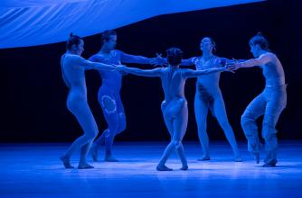 Five dancers are posed in a circle with their arms outstretched and touching, on a stage that is illuminated with blue lights