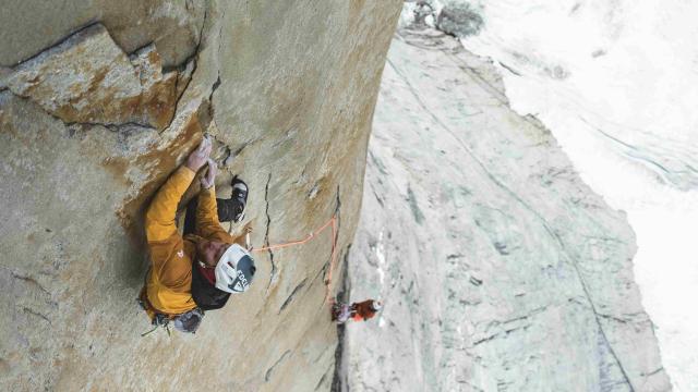 Man wearing yellow jacket climbing a big wall. Background shows a second man and glacier at the bottom. 