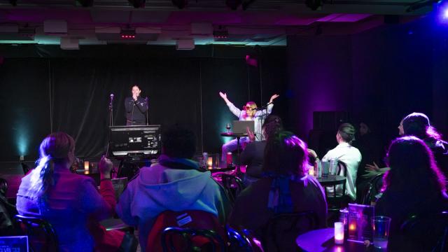 Audience seated at cabaret-style tables watches two performers on stage under colorful purple and green lighting during a live show.
