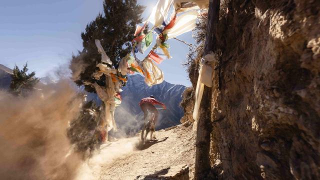 Biker speeding on a mountain road with Tibetan prayer flags hanging between trees and rocks