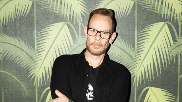 Portrait of a DJ standing confidently with arms crossed in front of a tropical palm leaf backdrop.