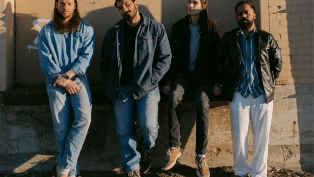 Four young men standing with a relax posture in front of a brick wall waring denim and 