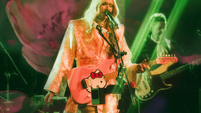 Performer with long platinum-blonde hair sings into a microphone while playing a pink electric guitar under green stage lights, with another musician performing in the background.