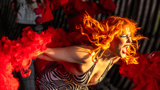 High-energy cabaret performance featuring a singer with vibrant red hair and dramatic red costume elements, surrounded by theatrical performers in an immersive stage setting.
