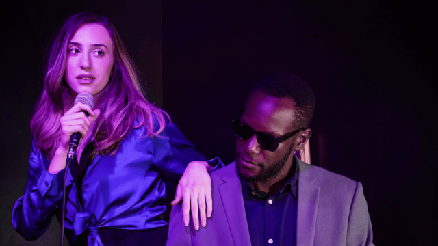 Singer holding a microphone stands beside a pianist wearing sunglasses under purple stage lighting during a live performance.