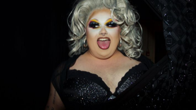 Performer wearing dramatic stage makeup, a platinum-blonde wig, and a black sequined outfit poses with their tongue out against a dark background.