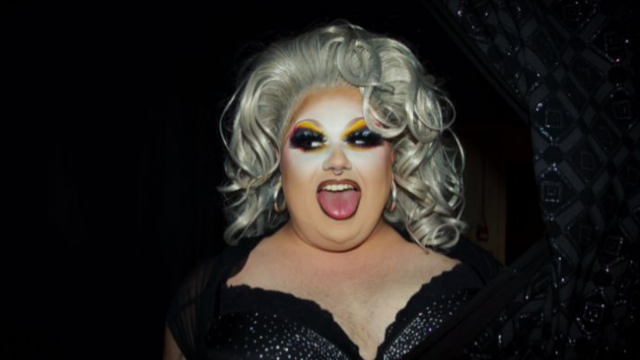 Performer wearing dramatic stage makeup, a platinum-blonde wig, and a black sequined outfit poses with their tongue out against a dark background.