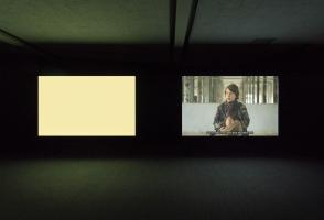 Two large screens in a dark room, one is a blank beige and the other vaguely shows a person.