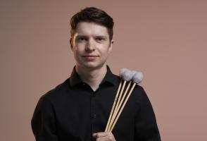 Nolan is pictured here in front of a red background holding mallets. 