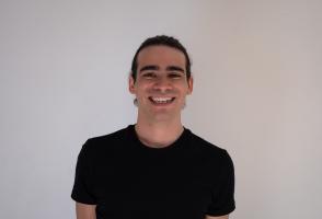 Sebastián stands in front of a white wall, smiling. He is wearing a black t-shirt. 