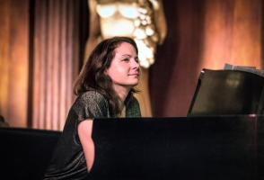Kris Davis sits at a black piano with pink curtains and a white statue behind her.