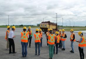 Photograph of twelve people wearing safety gear standing in a loose circle on a large concrete lot. There is a city garbage truck in the background.