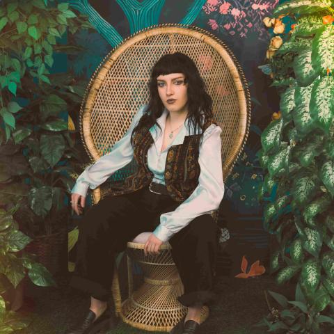 A person seated in a woven wicker chair surrounded by lush green plants and a colorful mural backdrop. They wear a white button-up shirt, patterned vest, black pants, and black shoes, with dark hair and dramatic makeup, posing confidently in a stylized indoor garden setting