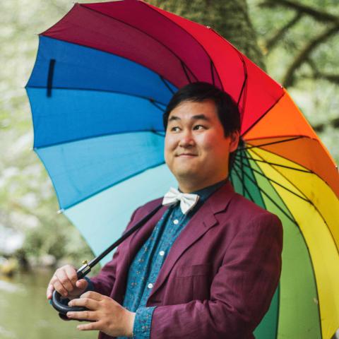 Man holding rainbow coloured umbrella open behind him and smiling offside to the camera