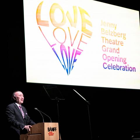 Jon stands behind a wooden podium with a rainbow heart projected on the screen behind him.