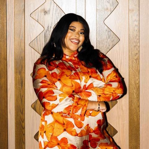 A smiling person with long dark hair poses confidently with crossed arms, wearing a flowing outfit covered in bold orange floral patterns. They stand against a light wood-paneled wall with geometric detailing, creating a warm and vibrant portrait