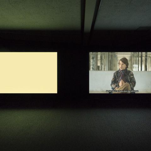 Two large screens in a dark room, one is a blank beige and the other vaguely shows a person.