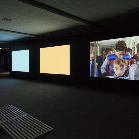 Three screens in a dark room, two are blank colours and the other shows three children.