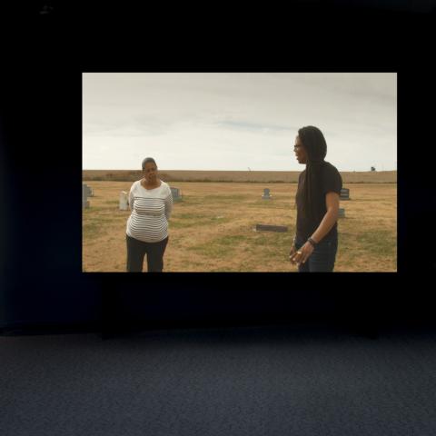 Large Screen in dark room depicting a lady in a white shirt speaking with a dark haired man.
