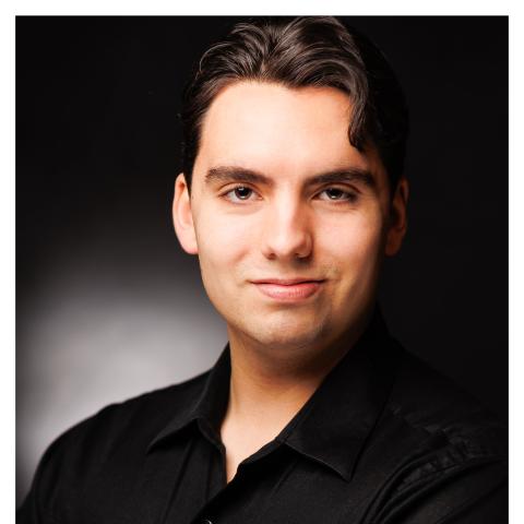 Man in black standing against dark background smiling lightly at camera with short wavy brown hair