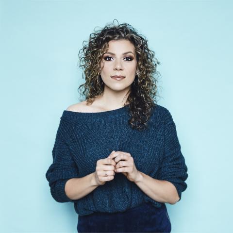 Alexis wears a dark dress in front of a blue background. She looks directly at the camera. Her hair is down.