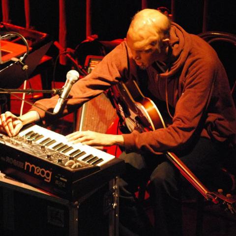 Shahzad Ismaily sits at a keyboard with a guitar in his lap. He is adjusting the keyboard and the photo is taken from ab