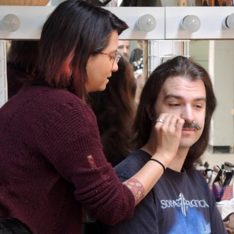 A long haired man gets a mustache applied by a woman