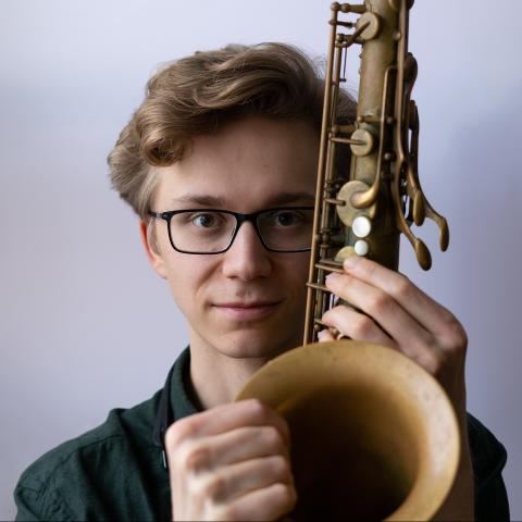 Young man wearing classes holding the saxophone bell facing front and to the side of his face