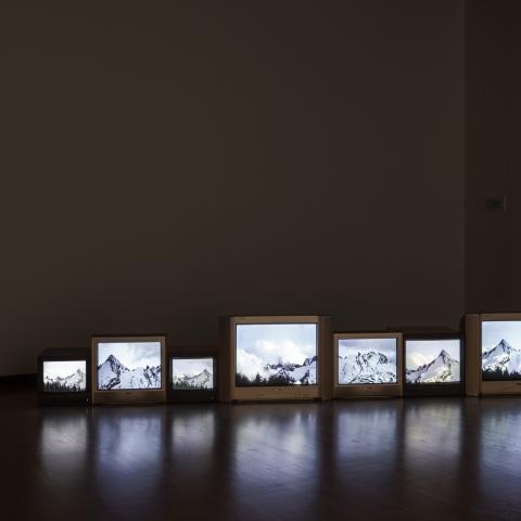 Seven TV screens of different sizes sit on a wooden floor. The TV screens all feature images of mountain ranges.